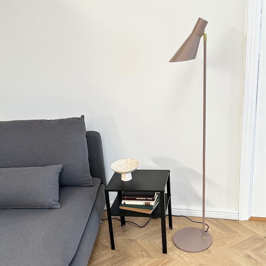 Floor lamp stands next to a small black side table; the table holds a decorative bowl and some books, all by a gray sofa in front of a white wall on a light wooden floor.