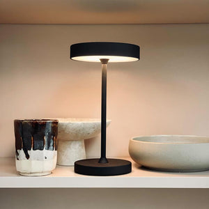 Table lamp with slim black stand casts soft warm light downwards; stands in the middle of a light shelf between a glazed ceramic cup and two muted stone-coloured bowls.