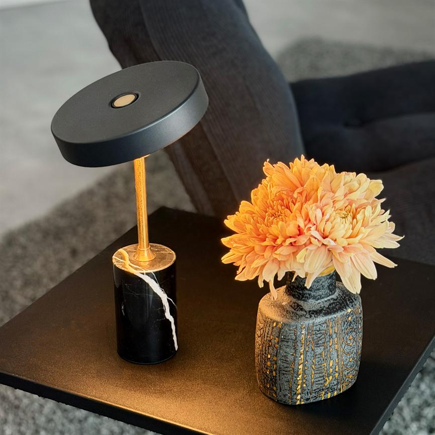 Table lamp casts warm light, next to a vase of peach-colored flowers, standing on a black coffee table in front of a dark sofa and a gray rug.