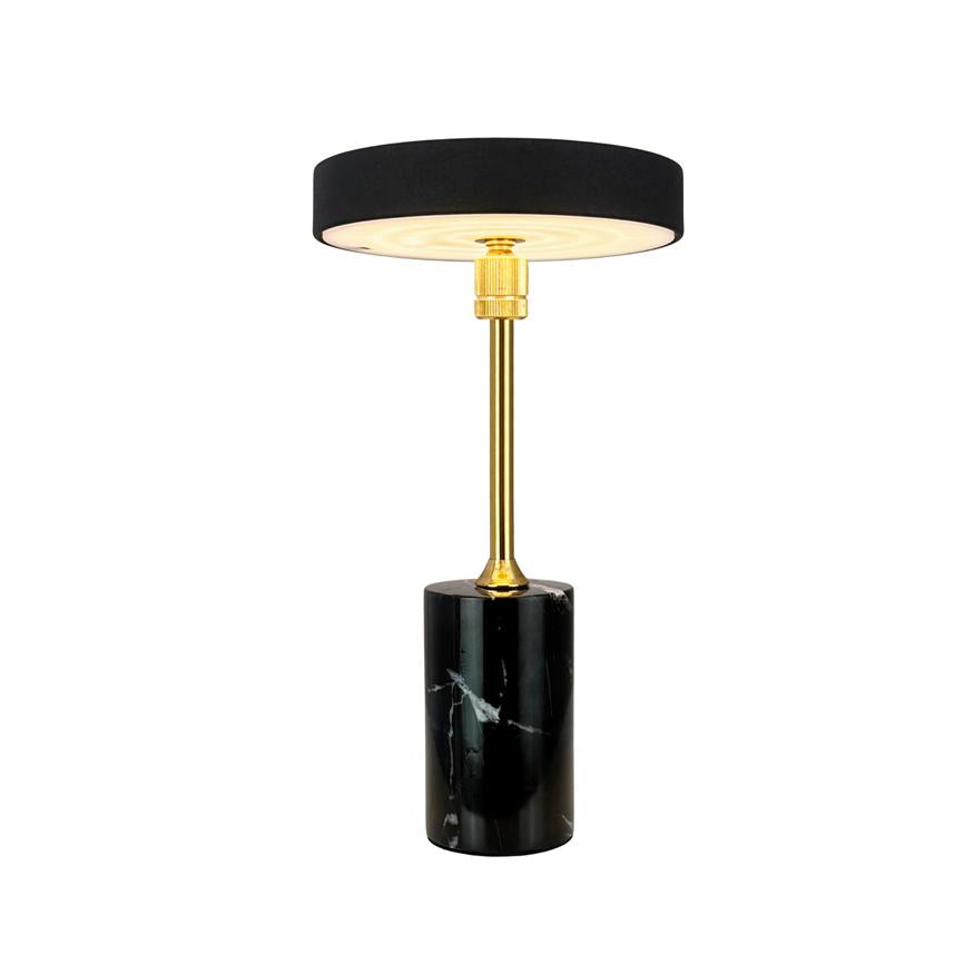 Table lamp with black round shade, golden rod and cylindrical black marble base; the lamp shines softly downwards and stands isolated against a white background.