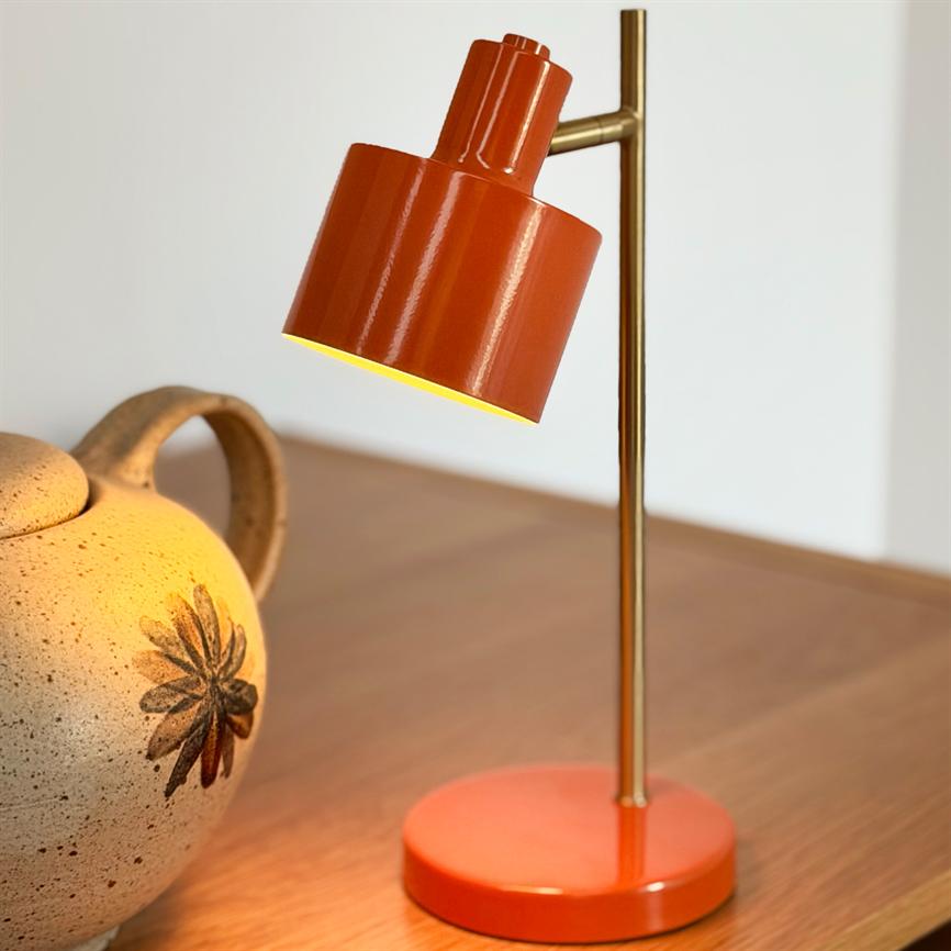 Red-orange table lamp casts warm light downwards; slender brass rod holds the screen above a wooden table next to a beige decorated ceramic jug.