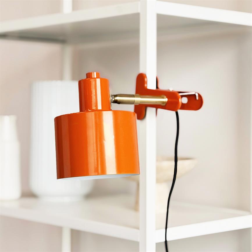 An orange metal wall lamp is clamped to a white bookcase and points downwards; a black cord hangs freely, surrounded by white vases and neutral decorations.