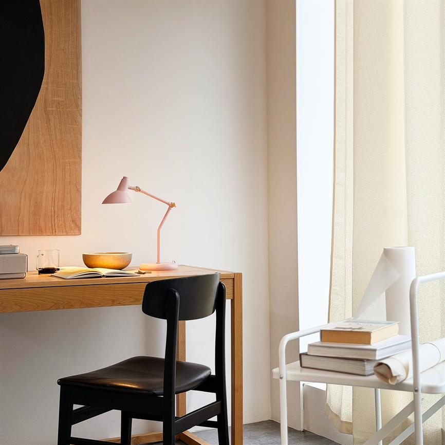 Pink desk lamp casts soft light over an open notebook and a bowl on a wooden desk; around is a black chair, light curtains and a white shelf with books.