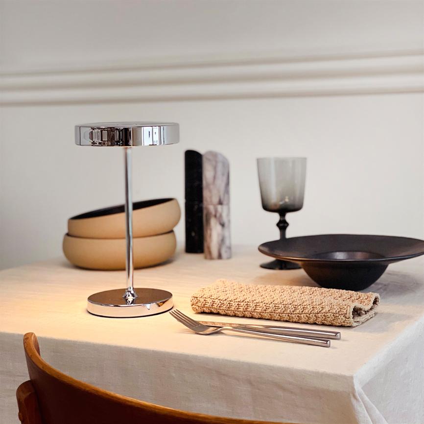 Chrome table lamp stands in the middle of a covered table; around it is a folded beige napkin, two forks, a black bowl, a smoked wine glass, ceramic bowls and marble stands.