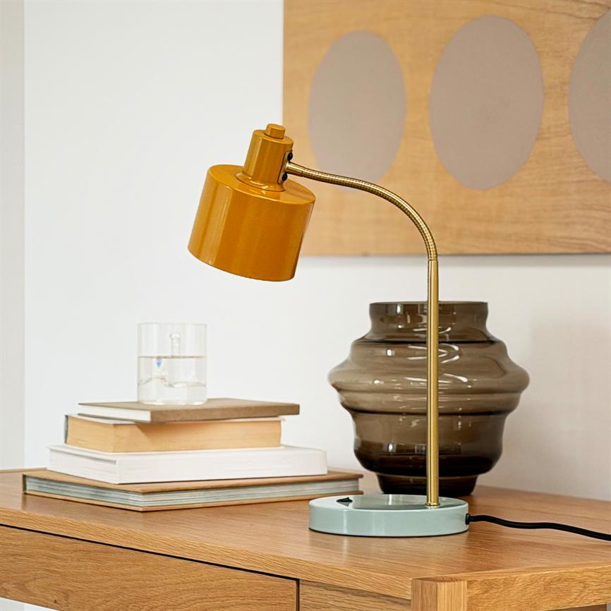 An ocher-colored table lamp with a curved gold-colored stem stands on a wooden table next to a stack of books and a smoke-colored glass vase in a bright, minimalist room with wall decoration.