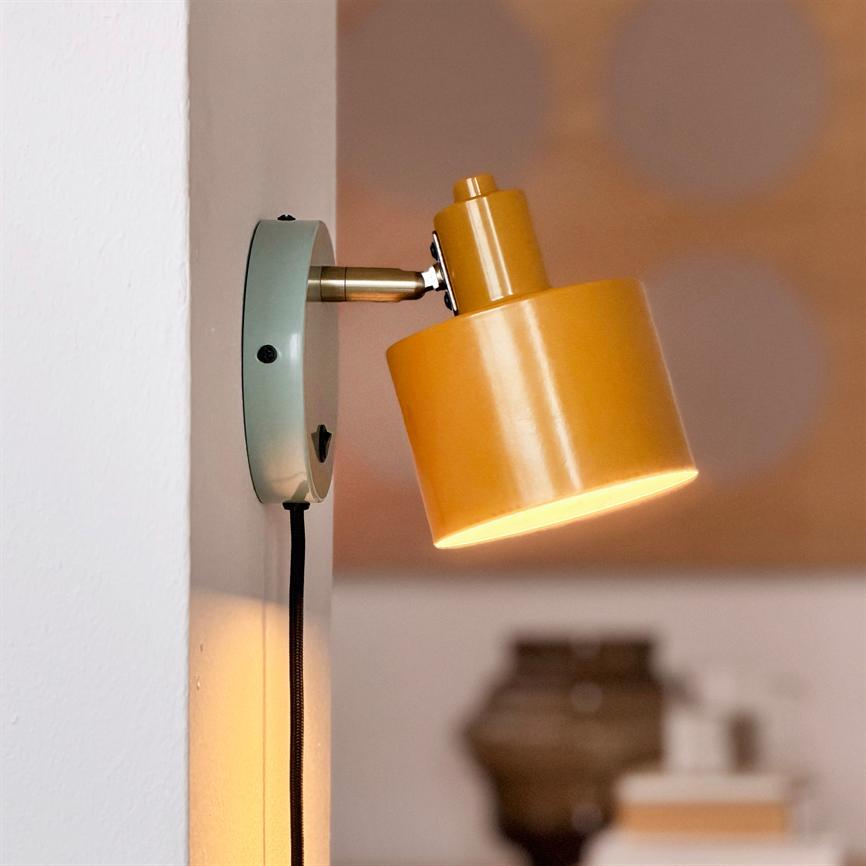 Yellow metal wall lamp faces downwards and casts warm light; mounted on round green wall bracket with black fabric cord, blurred indoor background.