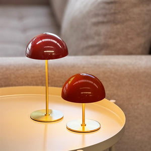 Two burgundy table lamps are lit on a round, bright metal table; low golden stands and hemispherical shades radiate warm light, surrounded by a beige sofa in the background.