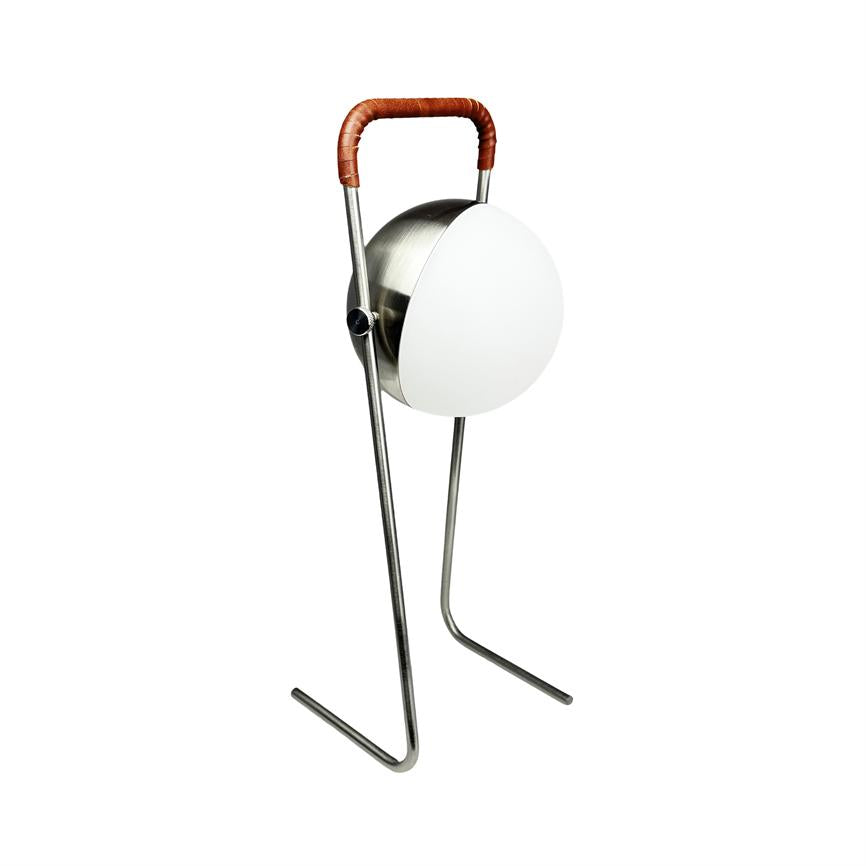 Lamp stands on two curved metal legs, with round white glass shade and round metal body, slightly angled; upper handle bound in brown leather. White, uniform background.