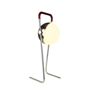 Floor lamp emits soft, warm light from a round, white shade; it stands on a slender chrome stand with two legs and a red leather-bound handle, photographed against a white background.