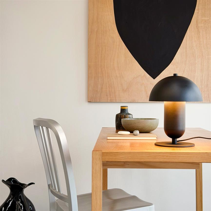 En mat sort bordlampe kaster varmt lys over et lyst træbord med keramikskål, flaske og bog; en metalstol står ved bordet, og et abstrakt maleri med sort form hænger på væggen.