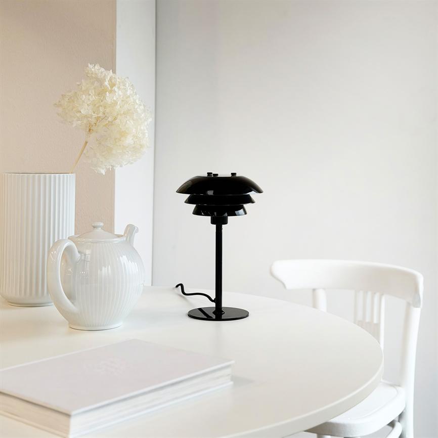 Sort bordlampe står på et rundt hvidt bord, omgivet af en hvid tekande, en høj ribbet vase med tørret blomst og en hvid stol foran en neutral væg.