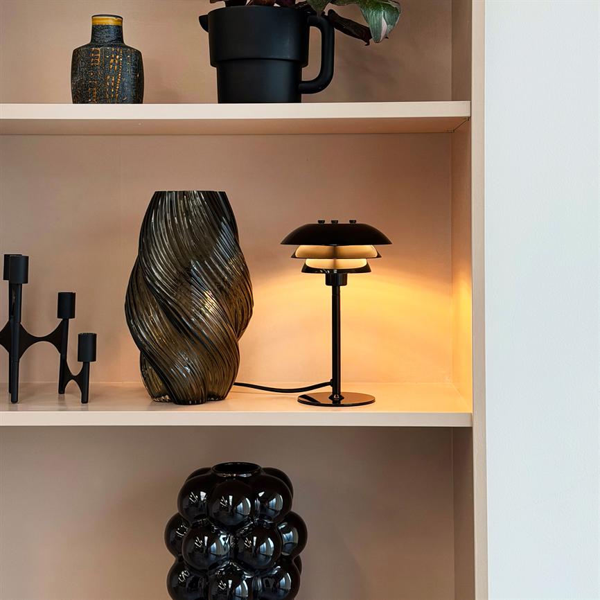 Sort bordlampe udsender varmt, gyldent lys på en hylde ved siden af en snoet røgfarvet glasvase og sorte dekorative genstande i en lys, indbygget reol.