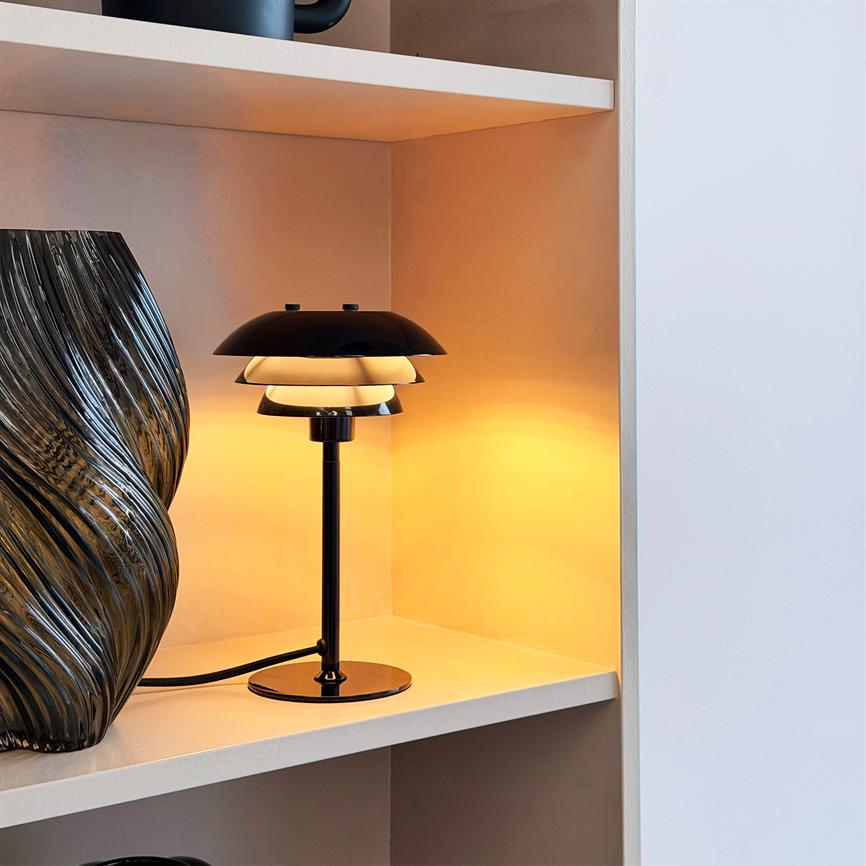Bordlampe udsender varmt, gyldent lys på en hvid hylde i en indbygget niche, ved siden af en mørk, ribbet glasvase; sort metalstamme og lagdelt skærm.