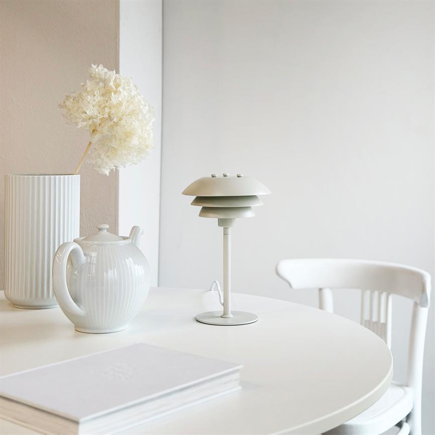 Bordlampen i lagdelt metal står slukket på et rundt hvidt bord, omgivet af en hvid tekande, en høj rilleret vase med tørret blomst og en hvid stol ved en lys væg.