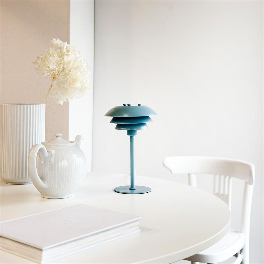 Lille turkis bordlampe står på et rundt hvidt bord; ved siden af står en hvid tekande og en høj rillede vase med tørret hortensia; enkelt, lyst skandinavisk rum.
