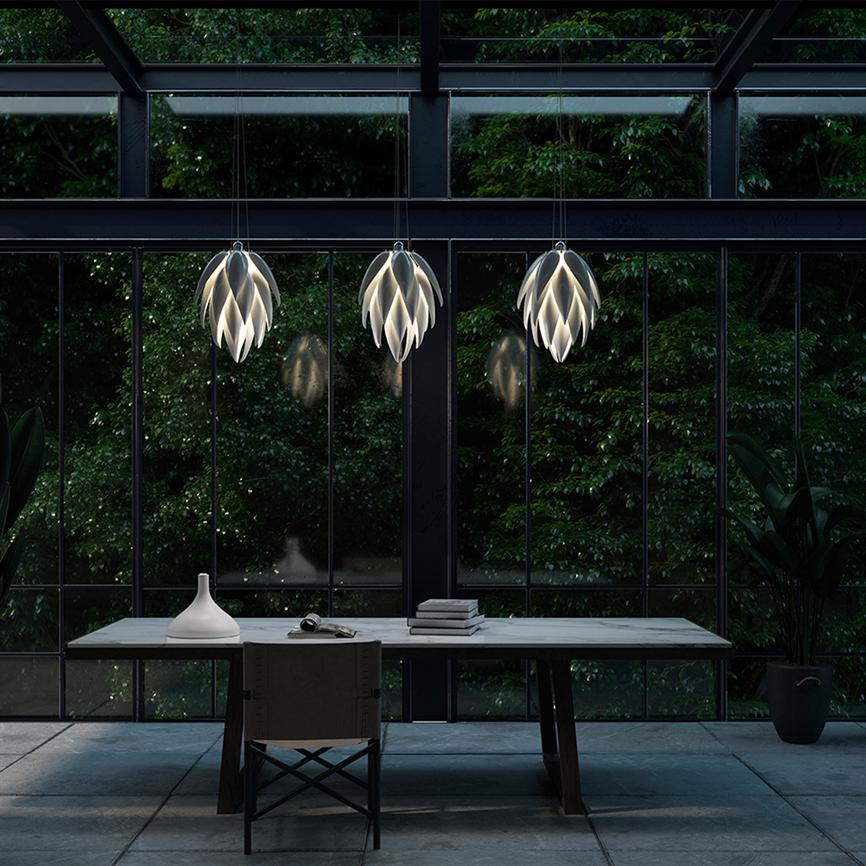 Three pendant lamps shaped like overlapping leaves hang and cast soft, warm light over a minimalist wooden table with chair, books and vase in front of large glass walls with dense, dark green planting outside.